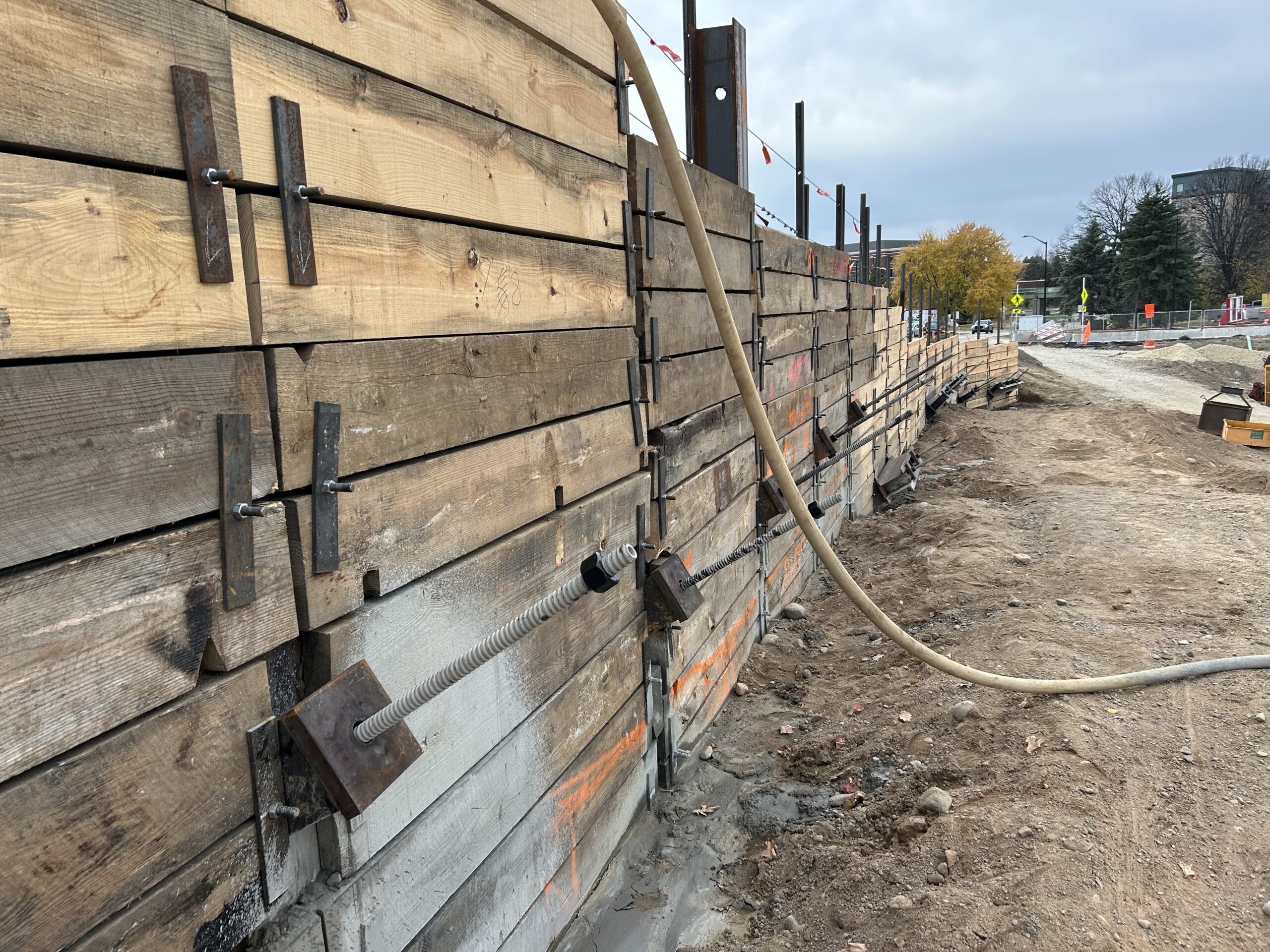 Wooden construction barrier with metal braces along a dug trench at a site, with hoses and dirt in the foreground.