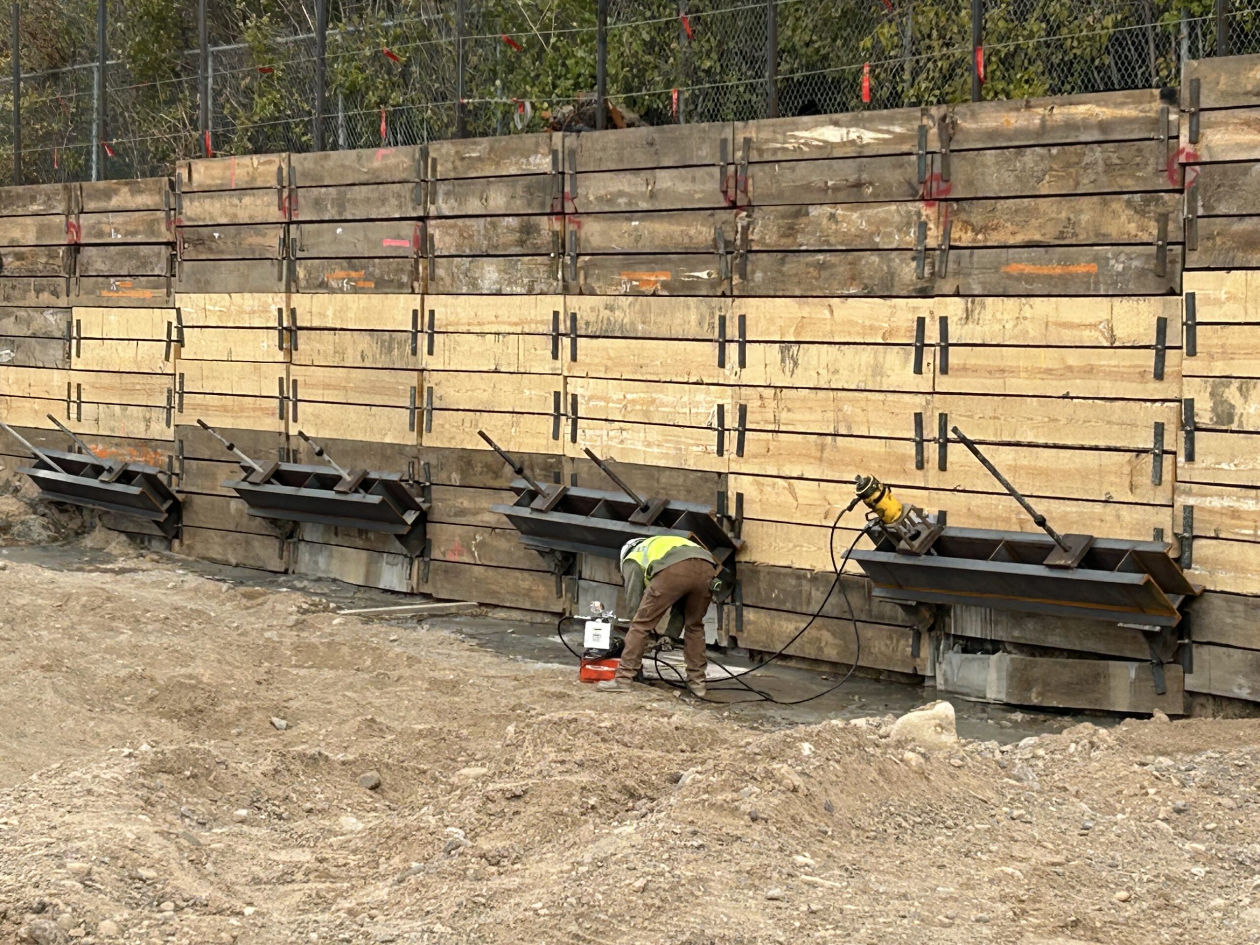 Construction site with workers assembling large wooden formwork panels for a concrete wall, using clamps and hoses visible in dirt foreground