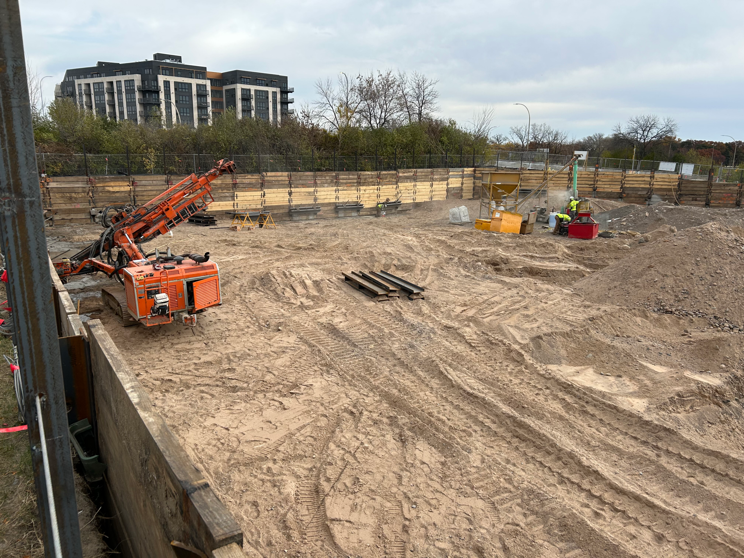 Construction site with an orange drilling rig in a deep excavation, workers in hi-vis vests, and wooden retaining walls; urban buildings in the background.