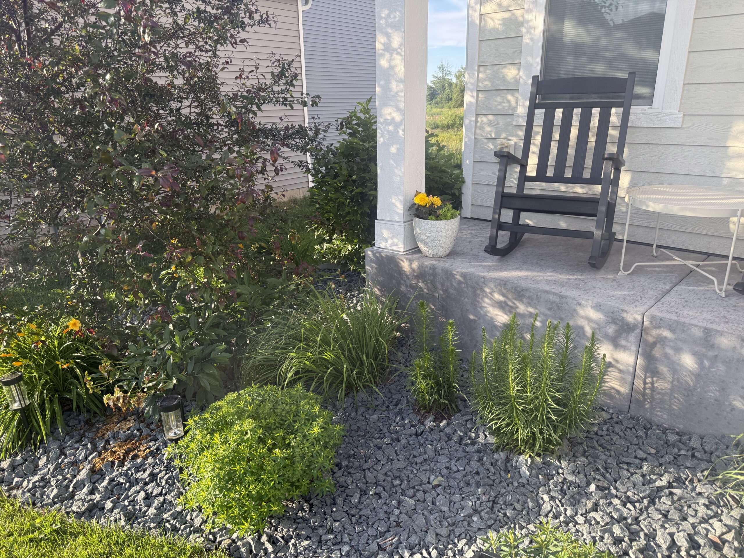 A gray rocking chair on a concrete porch beside a white planter with yellow flowers, near a window of a light-colored house.