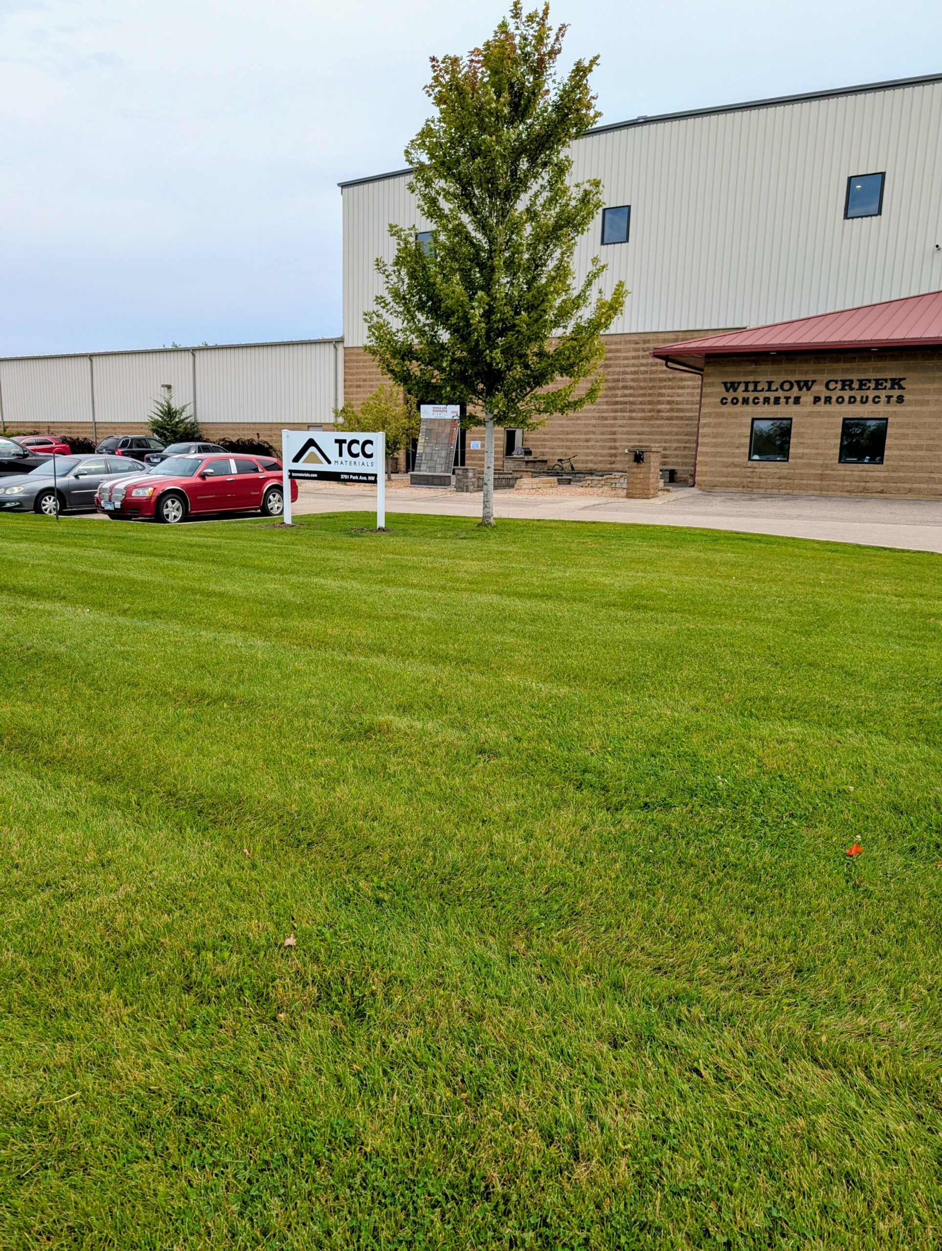 Front view of a commercial building with Willow Creek Concrete Products sign, a tree in front, and a row of parked cars nearby.