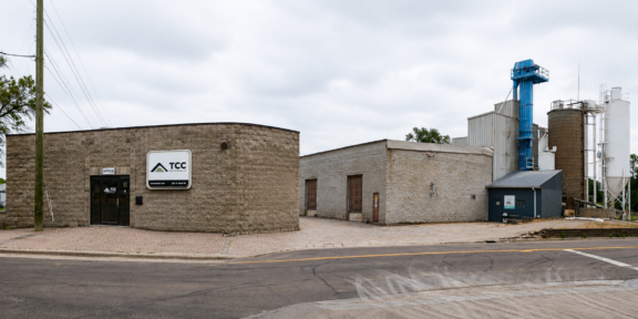 Industrial building with curved stone facade and a TCC Materials sign, adjacent warehouses and industrial silos in the background on a street.
