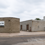 Industrial building with curved stone facade and a TCC Materials sign, adjacent warehouses and industrial silos in the background on a street.