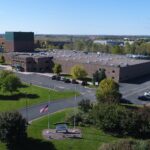 Aerial view of a large brick campus building with a tall green tower, surrounded by parking lots and manicured green lawns.