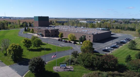 Aerial view of a large brick campus building with a tall green tower, surrounded by parking lots and manicured green lawns.
