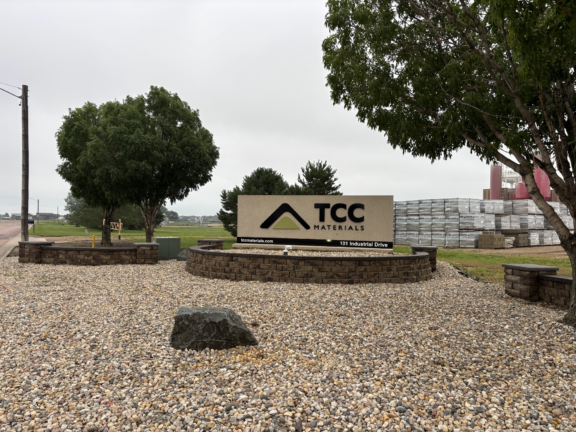 TCC Materials sign at a gravel entrance with trees and stacked building materials in the background.