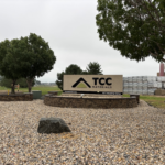 TCC Materials sign at a gravel entrance with trees and stacked building materials in the background.