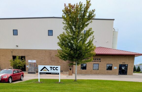 Commercial/industrial building with beige metal siding, brick base, and a large tree in front; TCC Materials sign near entrance.