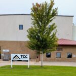 Commercial/industrial building with beige metal siding, brick base, and a large tree in front; TCC Materials sign near entrance.