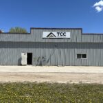 Front view of a gray corrugated-metal warehouse with a TCC Materials sign, set against a bright blue sky; yellow flowers cover the grassy foreground.