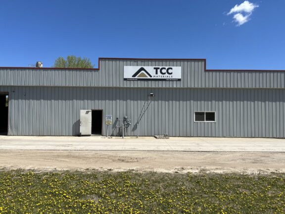 Front view of a gray corrugated-metal warehouse with a TCC Materials sign, set against a bright blue sky; yellow flowers cover the grassy foreground.