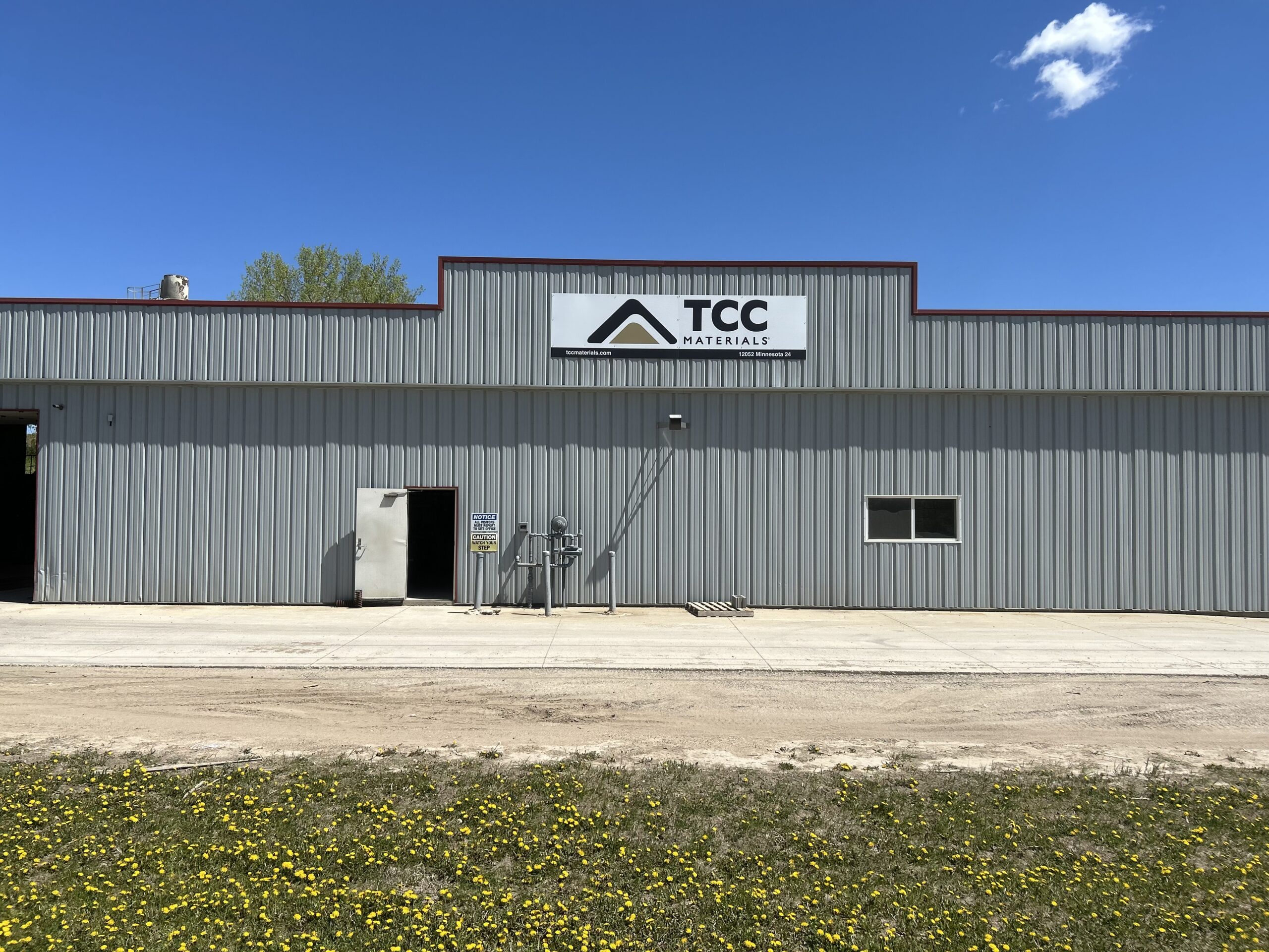 Front view of a gray corrugated-metal warehouse with a TCC Materials sign, set against a bright blue sky; yellow flowers cover the grassy foreground.