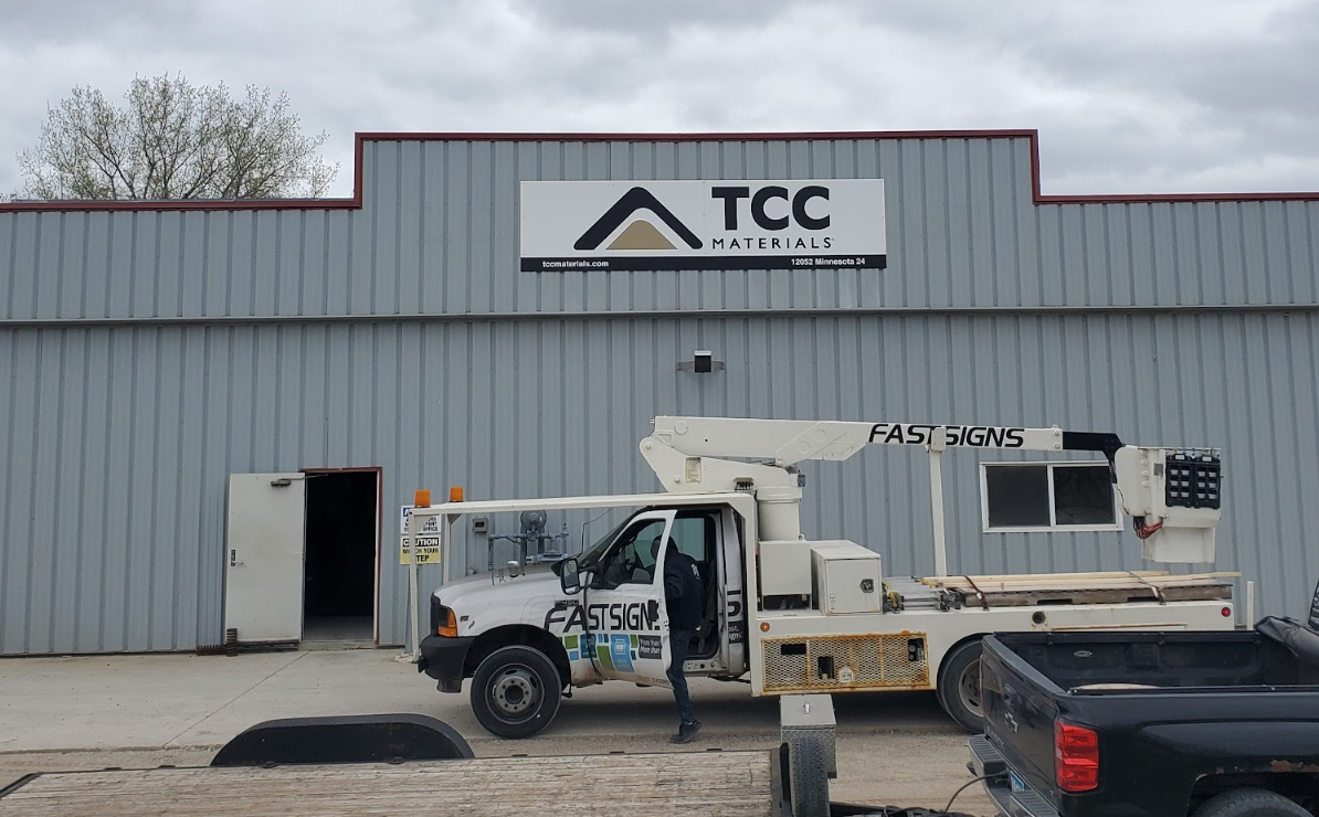 Exterior of a gray metal warehouse with a TCC Materials sign; a white bucket truck labeled FASTSIGNS is parked in front, with a person near the cab on a gray day.