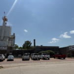 Industrial facility with a tall white tower, an American flag on top, and a row of white trucks parked in front under a clear blue sky.