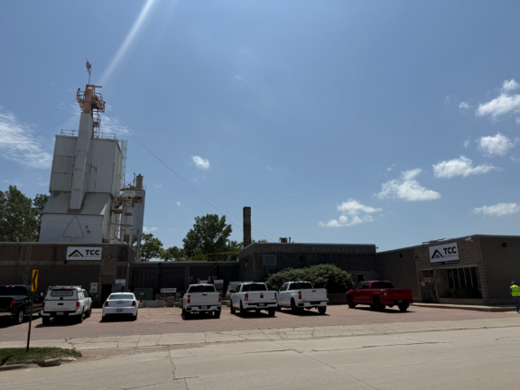 Industrial facility with a tall white tower, an American flag on top, and a row of white trucks parked in front under a clear blue sky.
