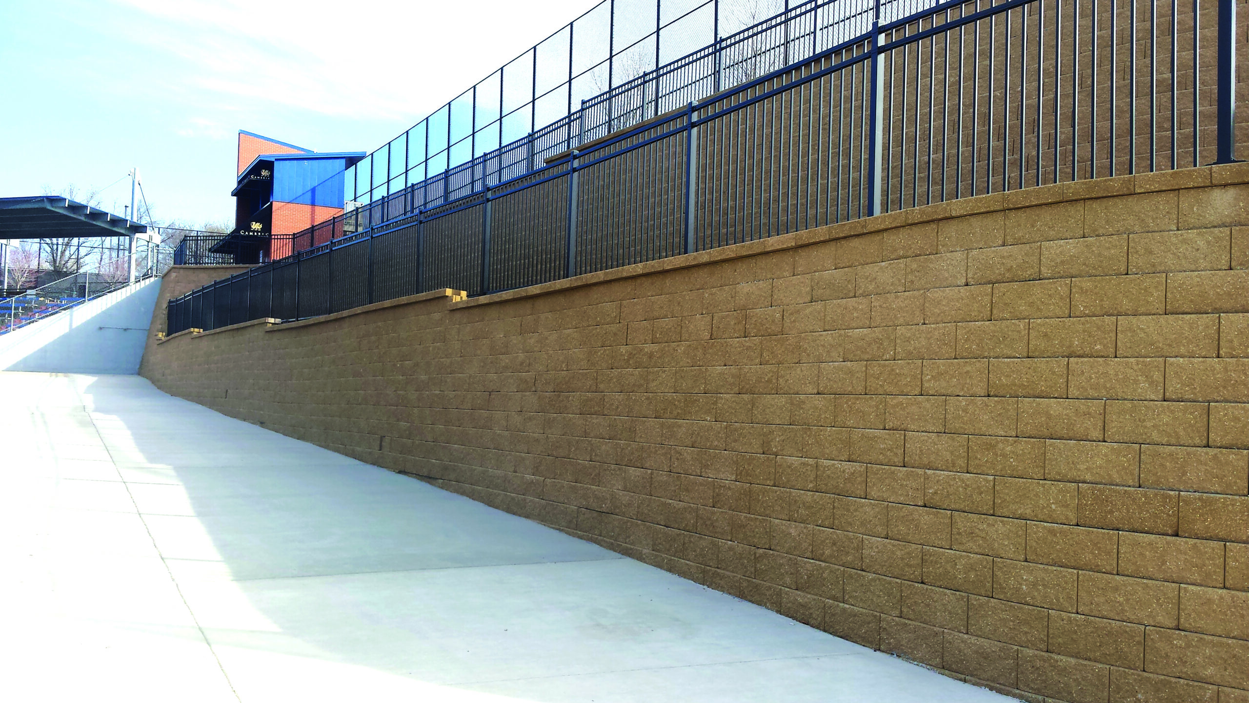 Sloped concrete walkway beside a tall brown brick retaining wall with a blue metal railing and a blue building in the distance.