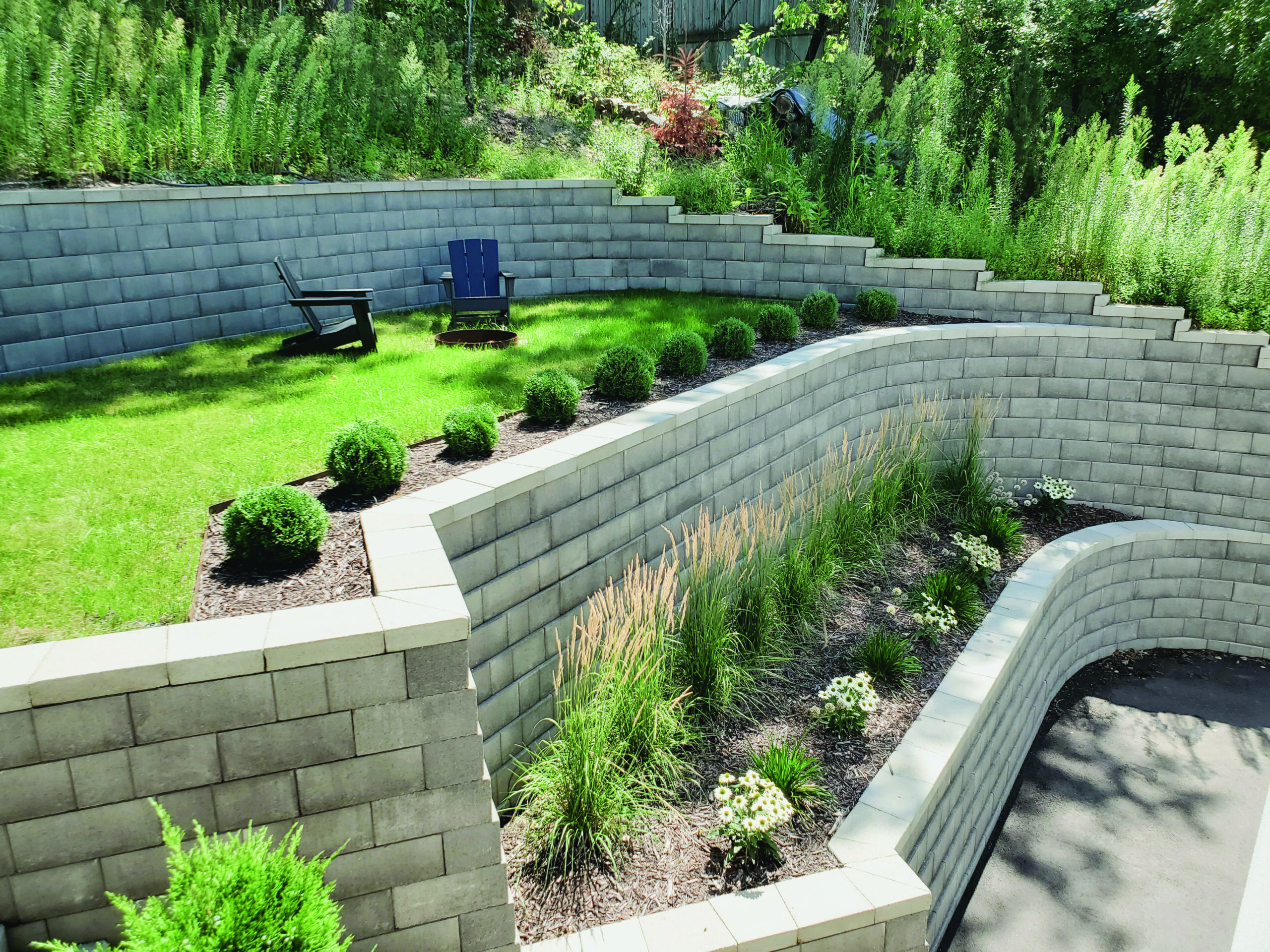 Tiered gray block retaining walls form a stepped garden with a small seating area and a circular fire pit on the lawn.