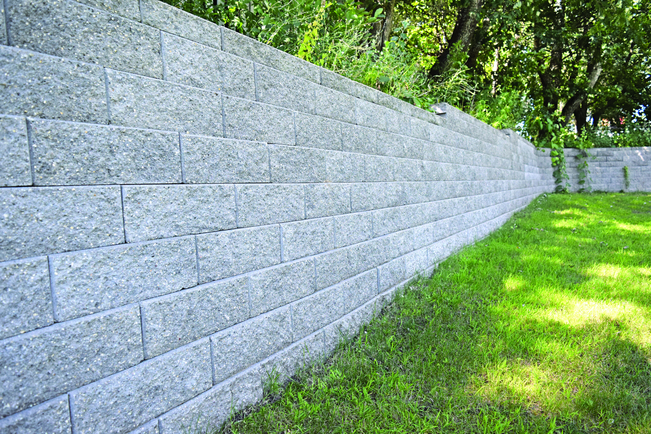 Long gray concrete block wall runs along a grassy lawn with trees visible behind it at the top edge.