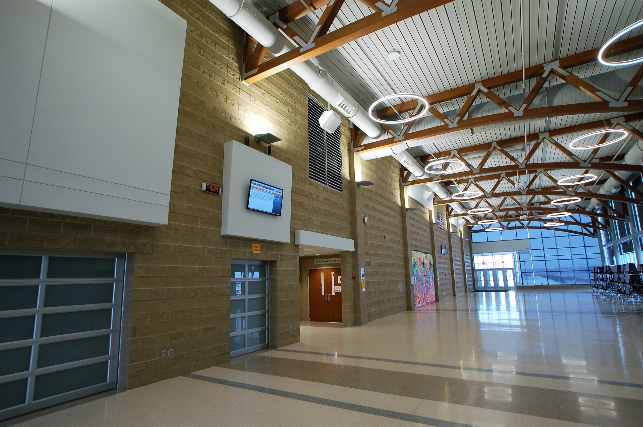 Wide indoor hallway in a modern building: brick walls, white panels, exposed wooden ceiling trusses, circular halo lights, and large windows at the far end of the hall.