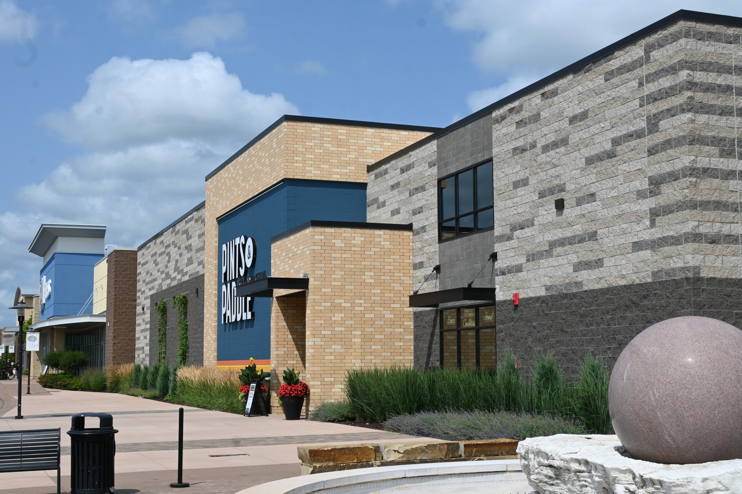 Exterior view of a modern retail center with tan brick and gray stone walls, a blue accent panel, and a large PINKS sign against a blue sky behind.