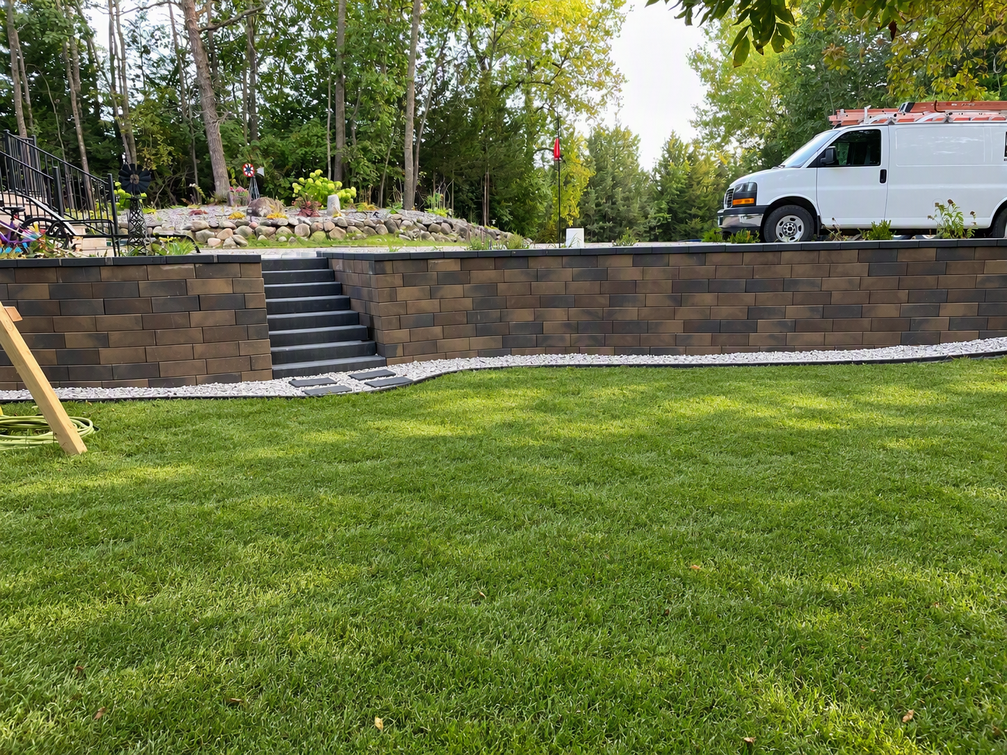 Brick retaining wall with stairs to an upper patio; a white van parked on the driveway beyond the wall in a wooded backyard scene.