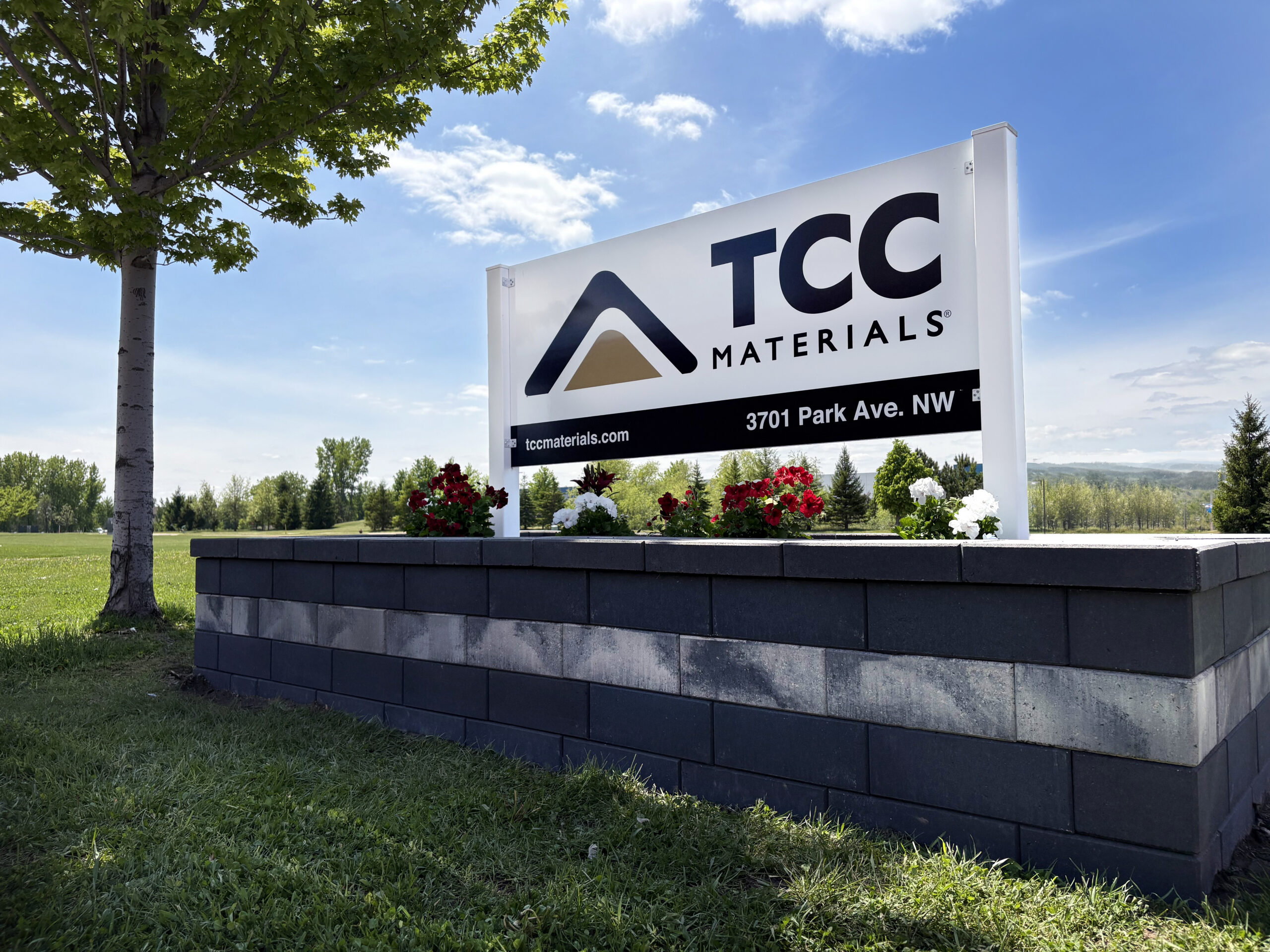 Outdoor TCC Materials sign with logo and address, set in a landscaped area with a flowering bed and blue sky above.