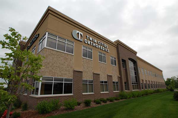 Exterior view of Twin Cities Orthopedics clinic building with a tan and brick facade and a well-kept lawn.