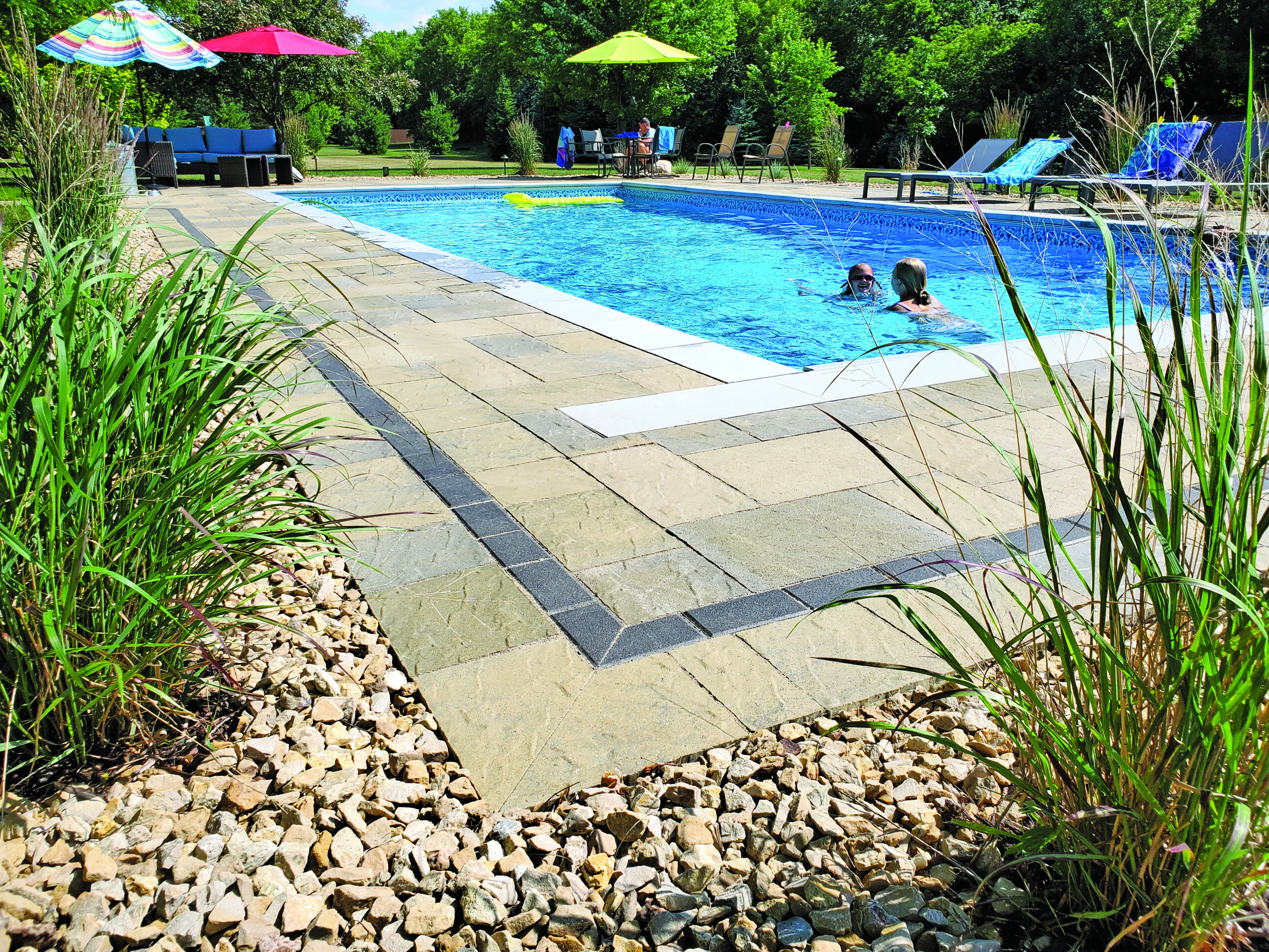 Two people swim in a bright blue rectangular backyard pool, surrounded by a stone deck and lounging chairs under colorful umbrellas.