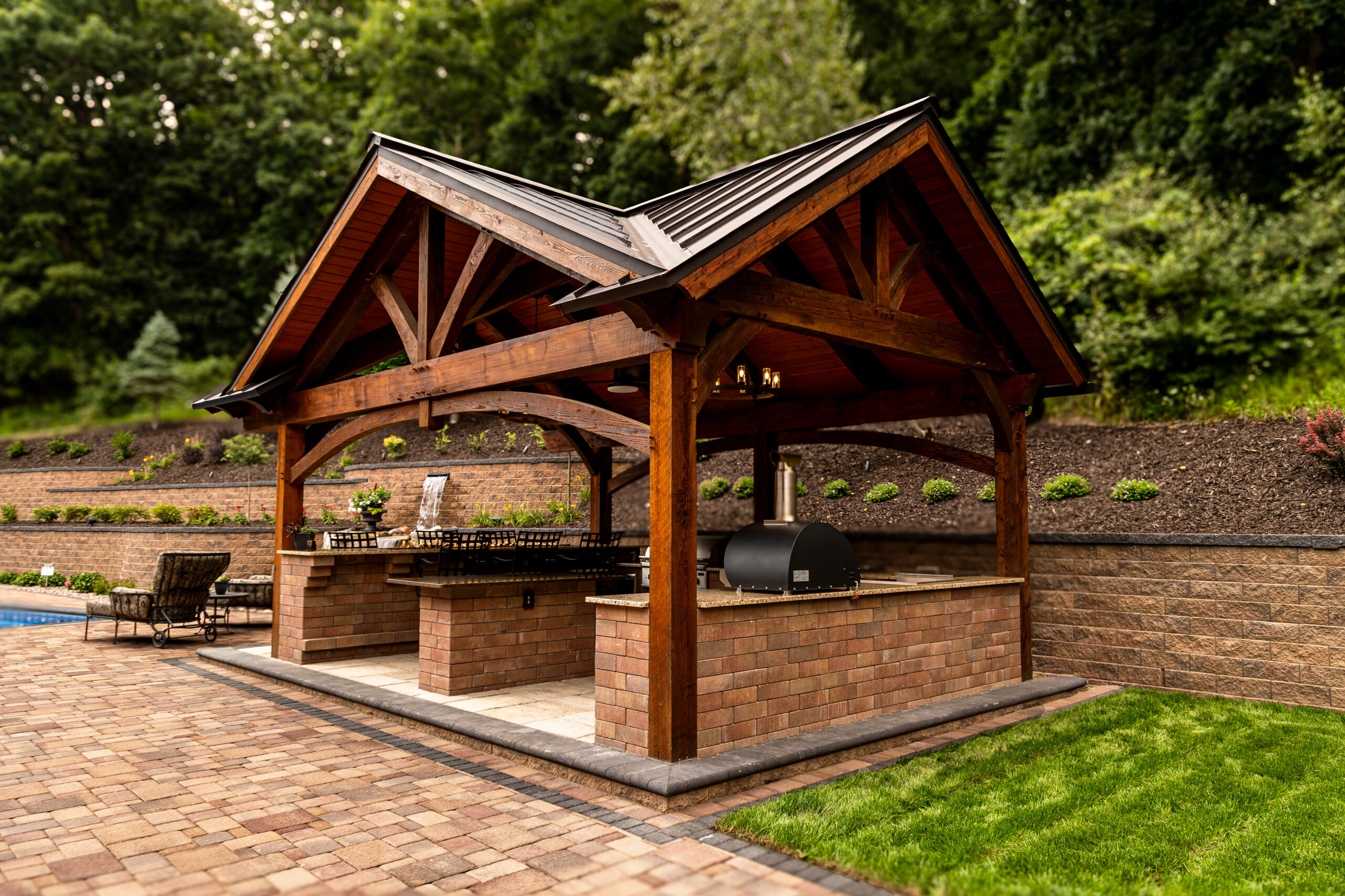 Wood-framed outdoor barbecue pavilion with brick counter, built-in grill, and landscaped yard behind it.