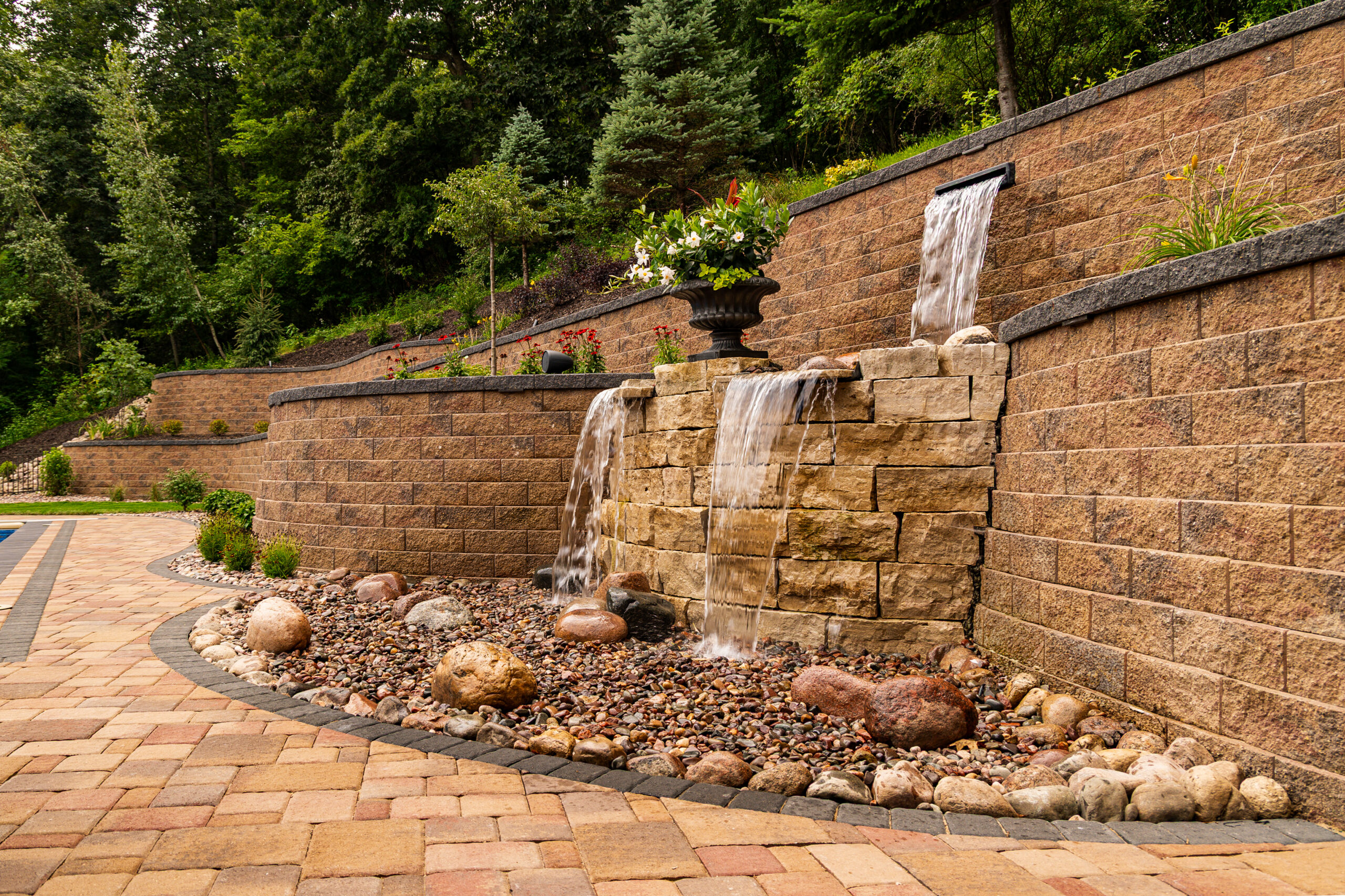 Tiered garden fountain set into a stone retaining wall, with water flowing down and a decorative planter on top amid shrubs and rocks.