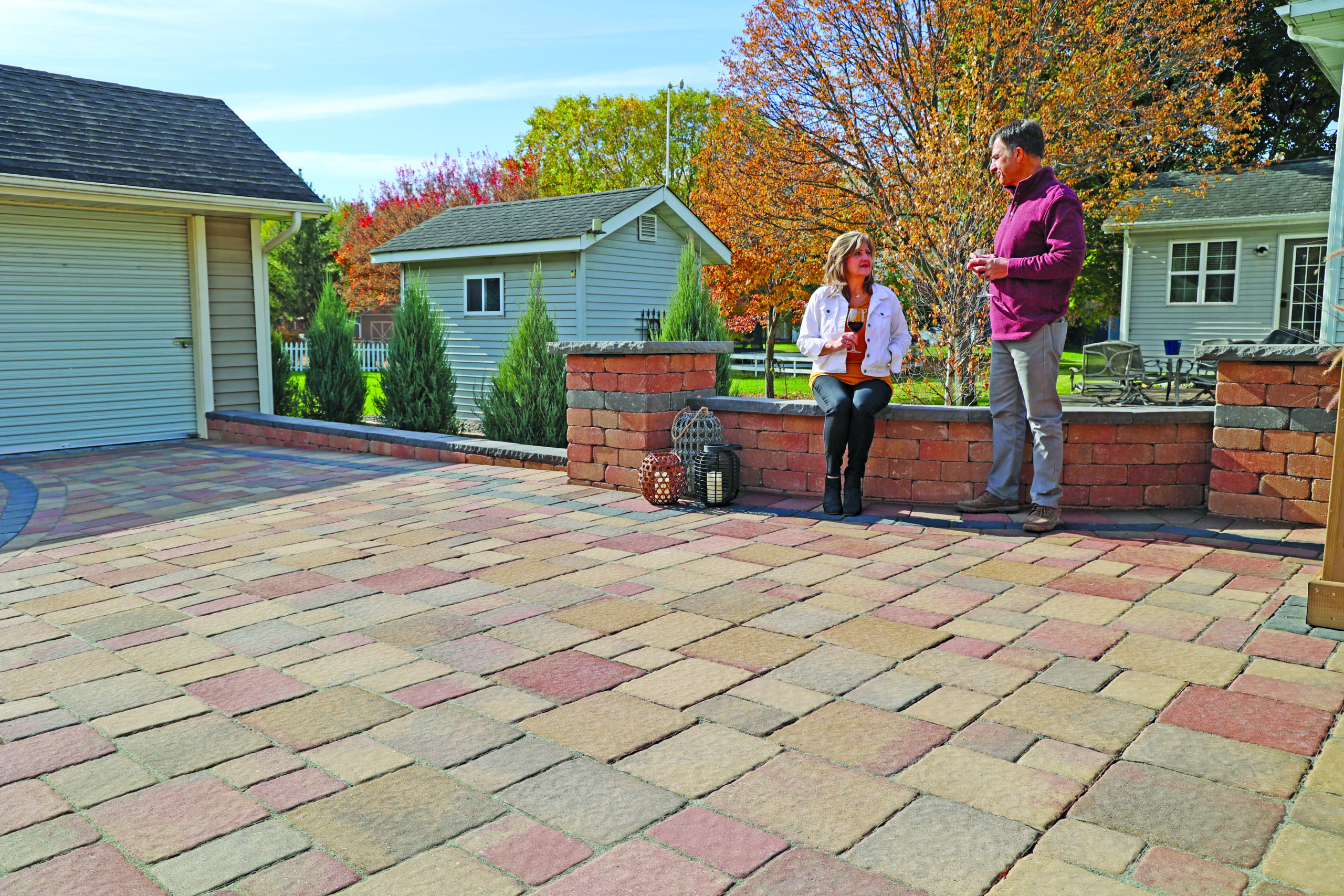 Man and woman standing on a brick patio chatting, autumn trees and houses behind them, sunny day.